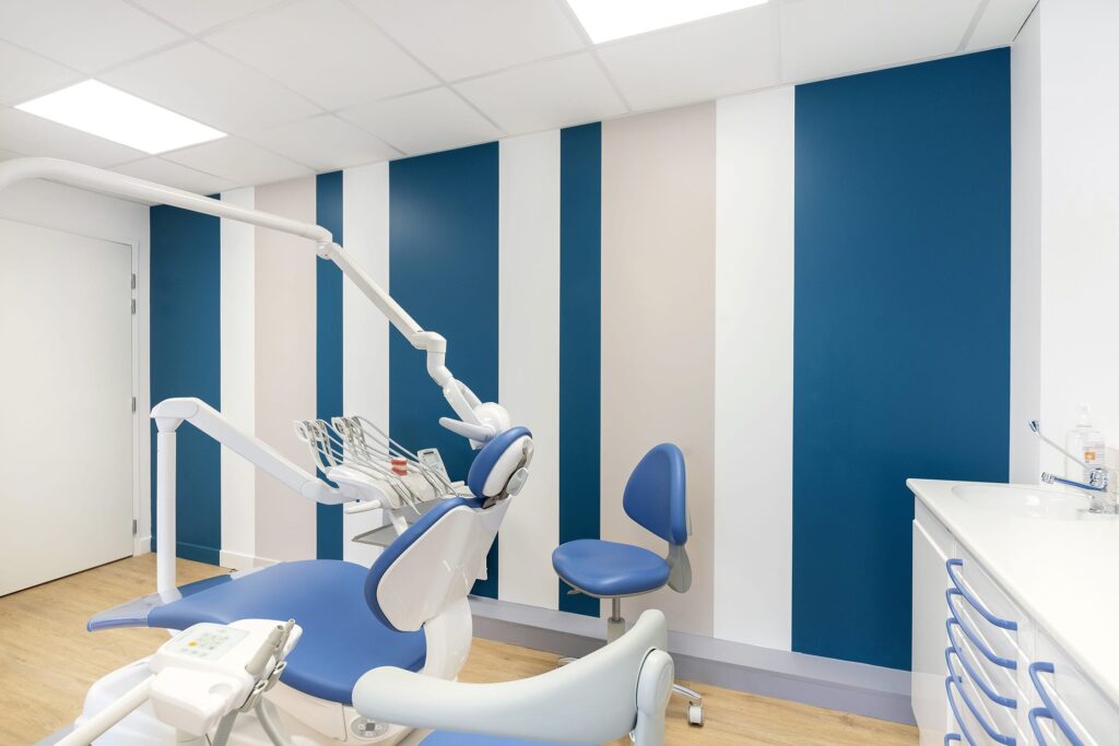 Salle de soins dentaires moderne avec murs rayés bleu et blanc, fauteuil dentaire, équipement et tiroirs de rangement ; intérieur clair et propre.