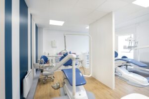 Intérieur d'une clinique dentaire moderne avec des chaises bleues et blanches, des équipements dentaires, des panneaux muraux rayés et un sol en bois. Deux postes dentaires sont séparés par une cloison partielle.