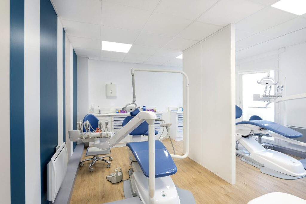 Clinique dentaire moderne au décor bleu et blanc, comprenant deux fauteuils dentaires, des équipements dentaires et des armoires sur un sol en bois clair.