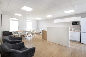 Cuisine de bureau lumineuse avec une longue table à manger, plusieurs chaises blanches, des fauteuils gris, des armoires blanches, un micro-ondes, un réfrigérateur et un sol en bois.