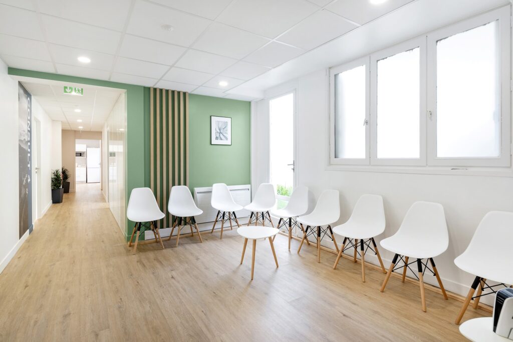 Une salle d'attente lumineuse avec des chaises blanches disposées en demi-cercle autour d'une petite table ronde, un sol en bois clair et des murs verts et blancs.