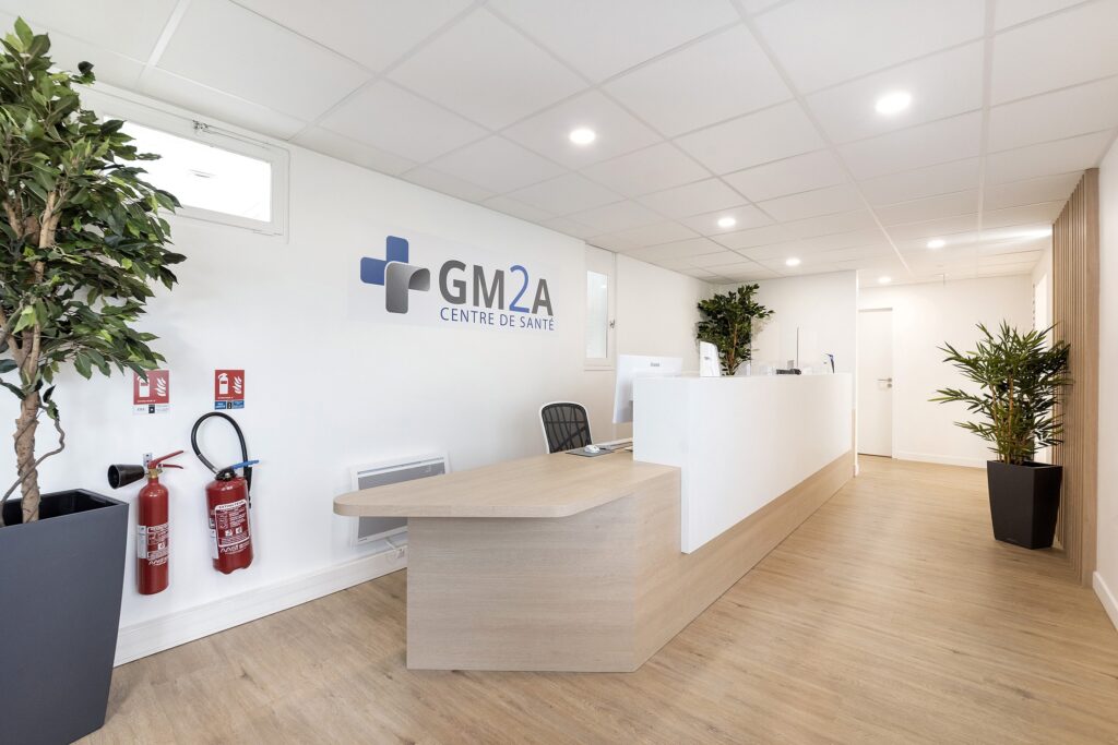 Réception d'un centre médical avec un bureau moderne, une chaise de bureau, des plantes en pot, des extincteurs et l'enseigne GM2A Centre de Santé sur le mur.