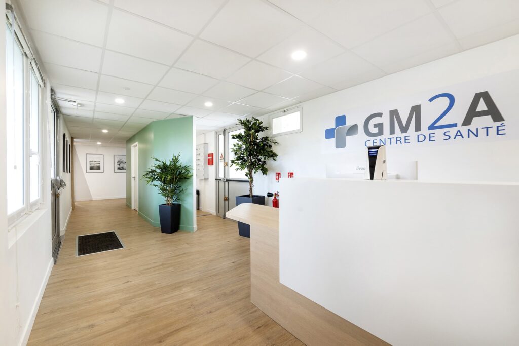 Réception d'un centre médical avec un bureau blanc, des plantes en pot, un sol en bois clair et l'enseigne "GM2A Centre de Santé" sur le mur.
