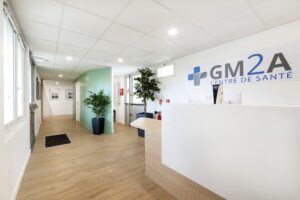 Réception d'un centre médical avec un bureau blanc, des plantes en pot, un sol en bois clair et l'enseigne "GM2A Centre de Santé" sur le mur.