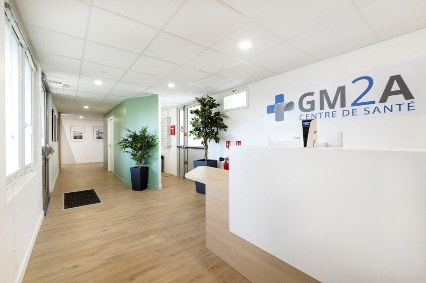 Réception d'une clinique médicale moderne avec un bureau blanc, des plantes vertes, un sol en bois et l'inscription "GM2A CENTRE DE SANTÉ" sur le mur.