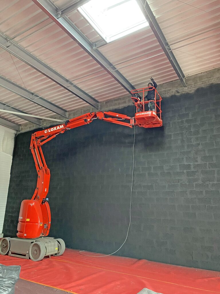 Une personne utilise un élévateur à nacelle rouge pour peindre ou travailler près du sommet d'un grand mur de briques noires à l'intérieur d'un entrepôt.