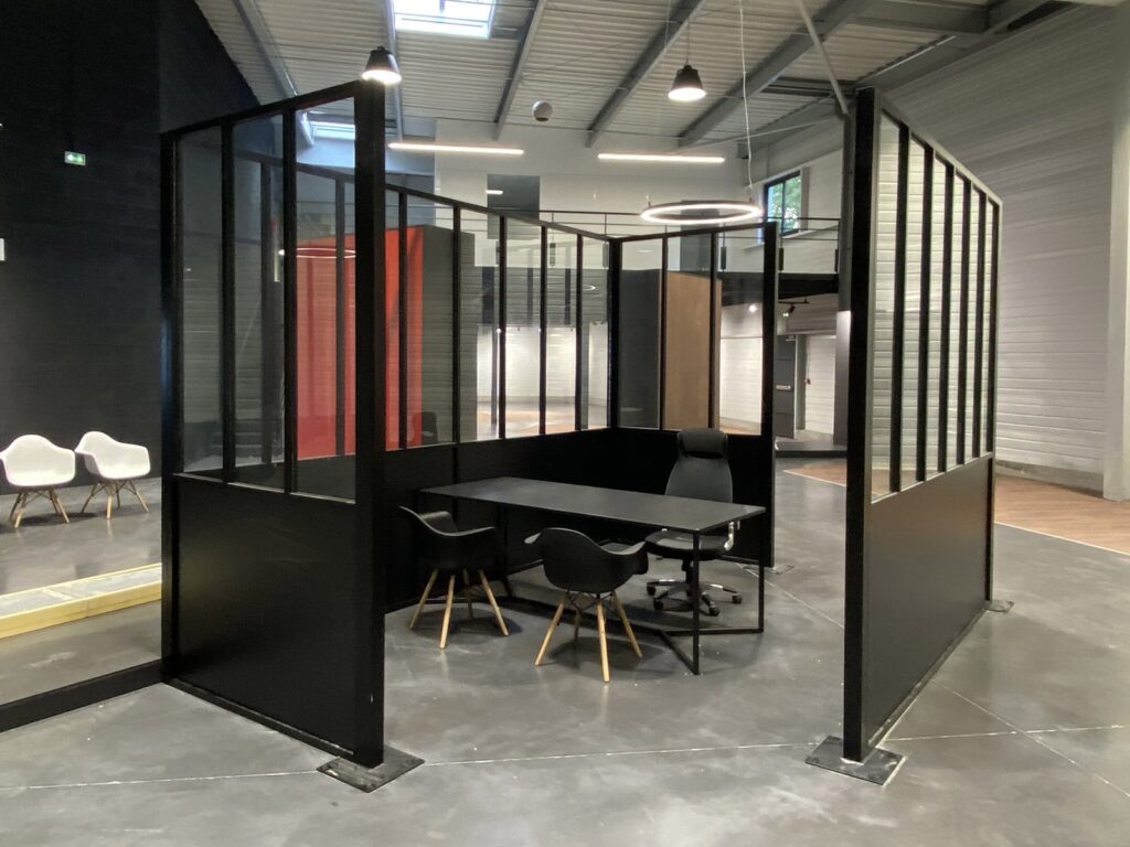 Bureau moderne avec des panneaux en verre et en métal noir, contenant une table, quatre chaises et un plafonnier circulaire dans un bâtiment de style industriel.