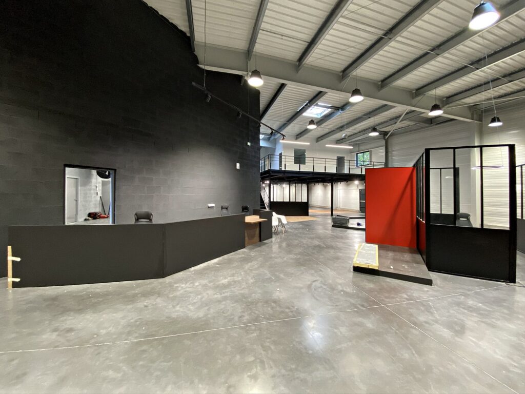 Un intérieur de bureau industriel spacieux avec un bureau d'accueil noir, un mur d'accent rouge, un sol en béton et une mezzanine en métal sous des plafonniers exposés.