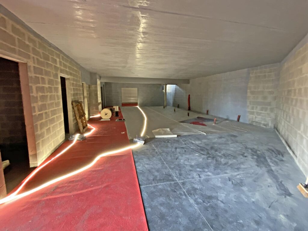 Espace intérieur inachevé avec des murs en blocs de béton, une moquette rouge partiellement posée, des câbles apparents, des matériaux de construction et des sections de plancher en cours de réalisation.