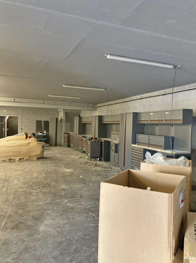 Une grande pièce inachevée avec des cartons, des outils et des armoires de rangement éparpillés le long des murs ; des plafonniers sont suspendus au-dessus de la tête.