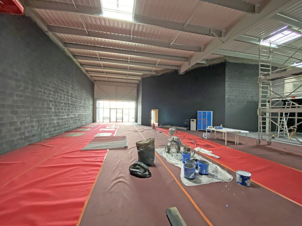 Une grande salle industrielle avec des revêtements de sol rouges, des murs en briques peintes en noir, du matériel de peinture et des échafaudages, éclairée par des puits de lumière et une grande porte vitrée à l'extrémité.