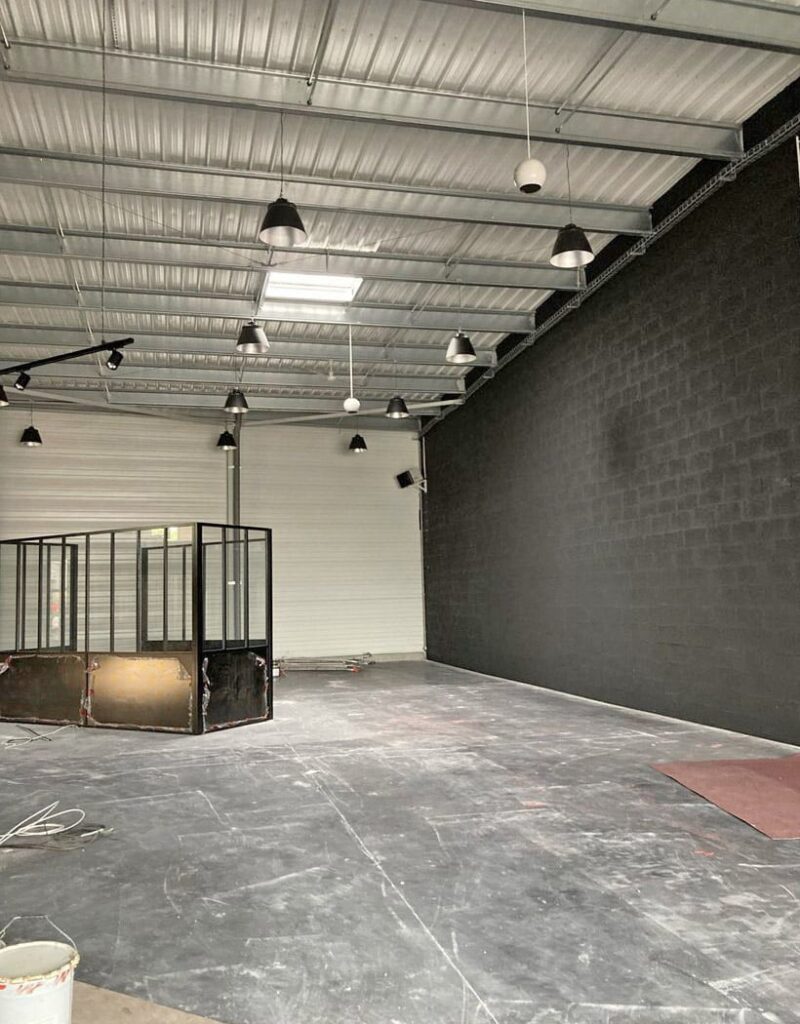Espace industriel vide avec un mur de briques noires, un toit en métal, plusieurs lampes suspendues et une enceinte en verre et en métal.