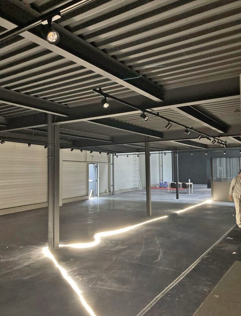 Intérieur d'un bâtiment de style industriel avec des poutres métalliques apparentes, un sol noir et une bande lumineuse qui court le long du sol ; une personne se tient sur le bord droit.