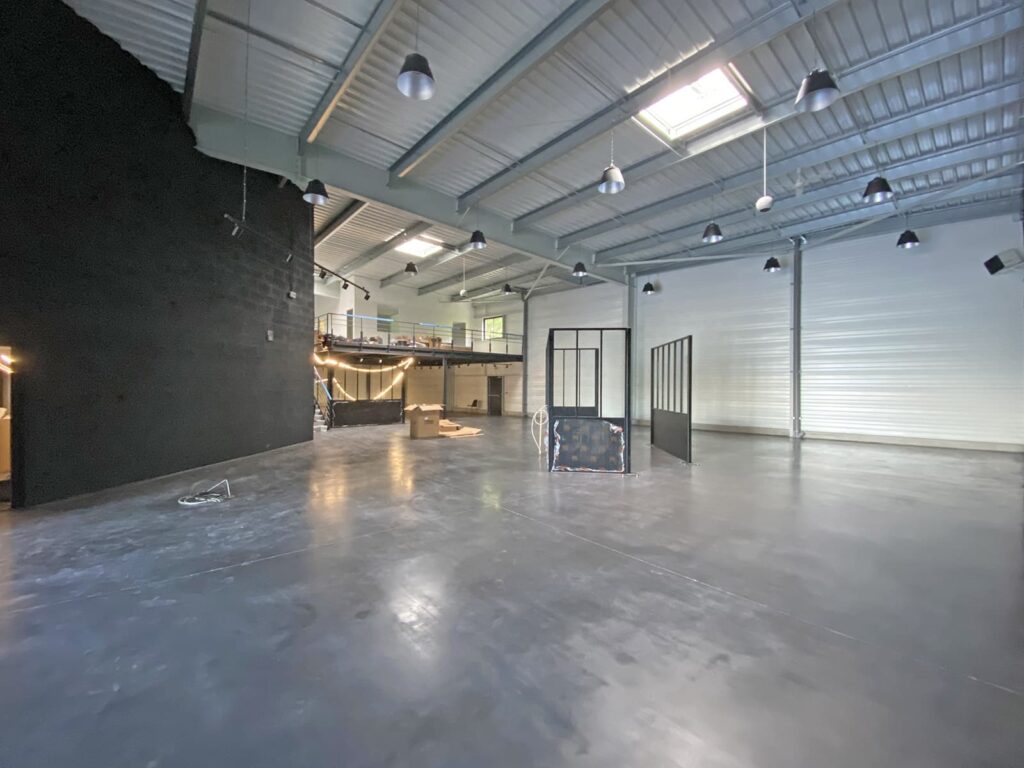 Intérieur industriel spacieux avec sol en béton, haut plafond, murs noirs et blancs, lampes suspendues et un espace loft avec des cloisons en verre.