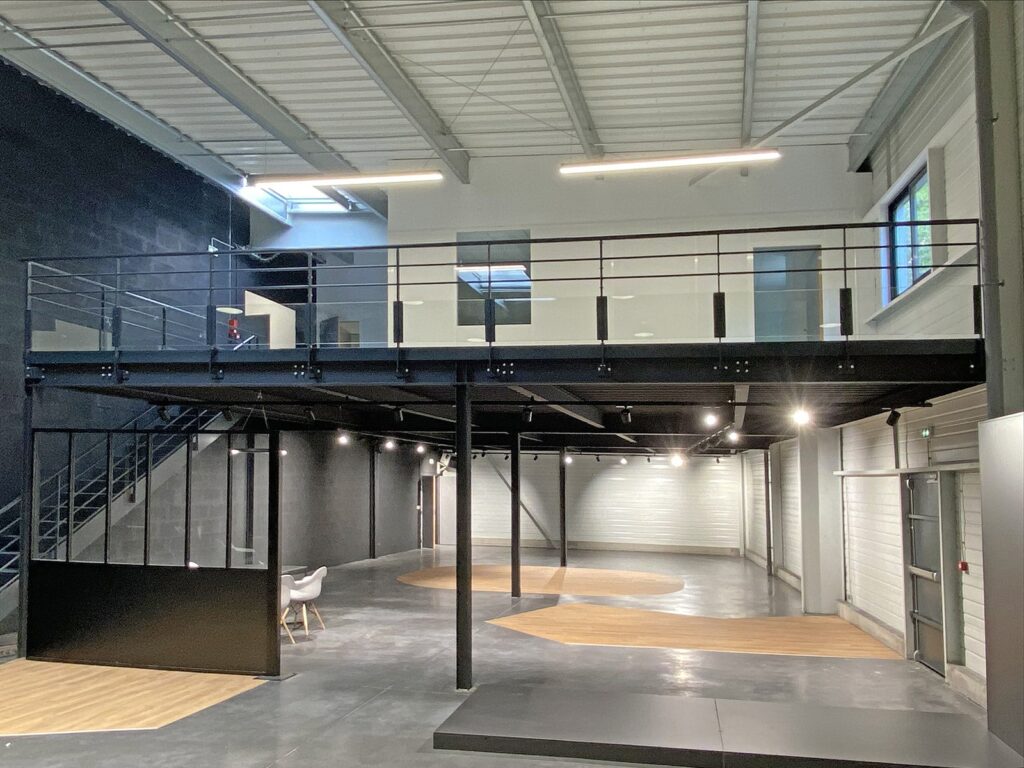 Intérieur industriel moderne et spacieux comprenant une mezzanine, des garde-corps métalliques, de grandes fenêtres et un sol en béton poli avec des accents en bois.