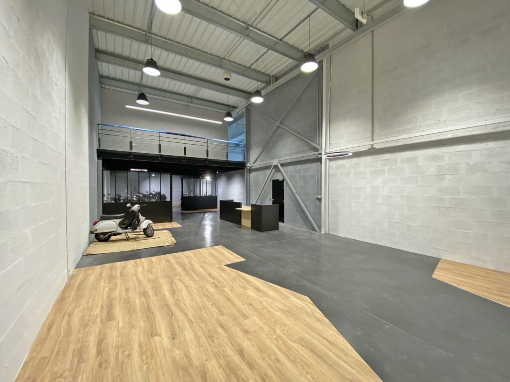 Un intérieur spacieux de style industriel avec de hauts plafonds, des murs gris, un sol en bois et en béton, une exposition de scooters et une mezzanine avec une rampe en verre.