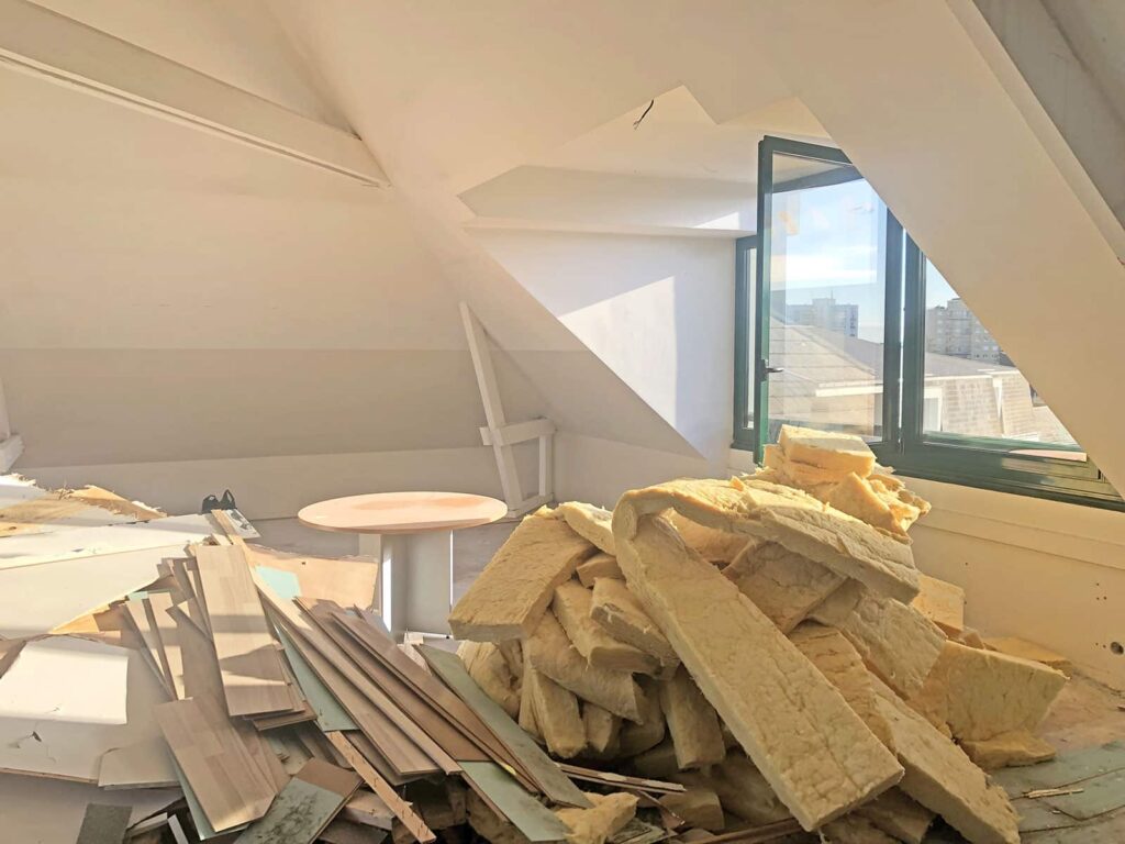 Chambre mansardée avec des matériaux d'isolation et des débris de construction éparpillés sur le sol, la lumière du soleil passant par une fenêtre ouverte.