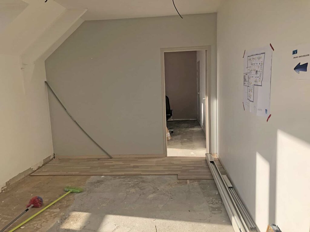 Pièce partiellement rénovée avec un sol inachevé, des outils et des matériaux éparpillés, et une porte menant à une autre zone ; un plan est collé sur le mur de droite.