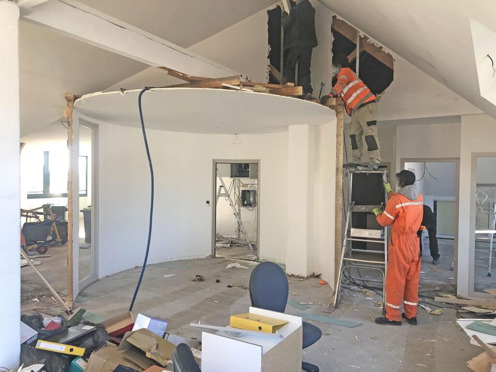 Deux ouvriers en tenue de sécurité enlèvent une partie d'un mur intérieur dans un bureau en cours de rénovation, avec des débris et du mobilier de bureau éparpillés autour.