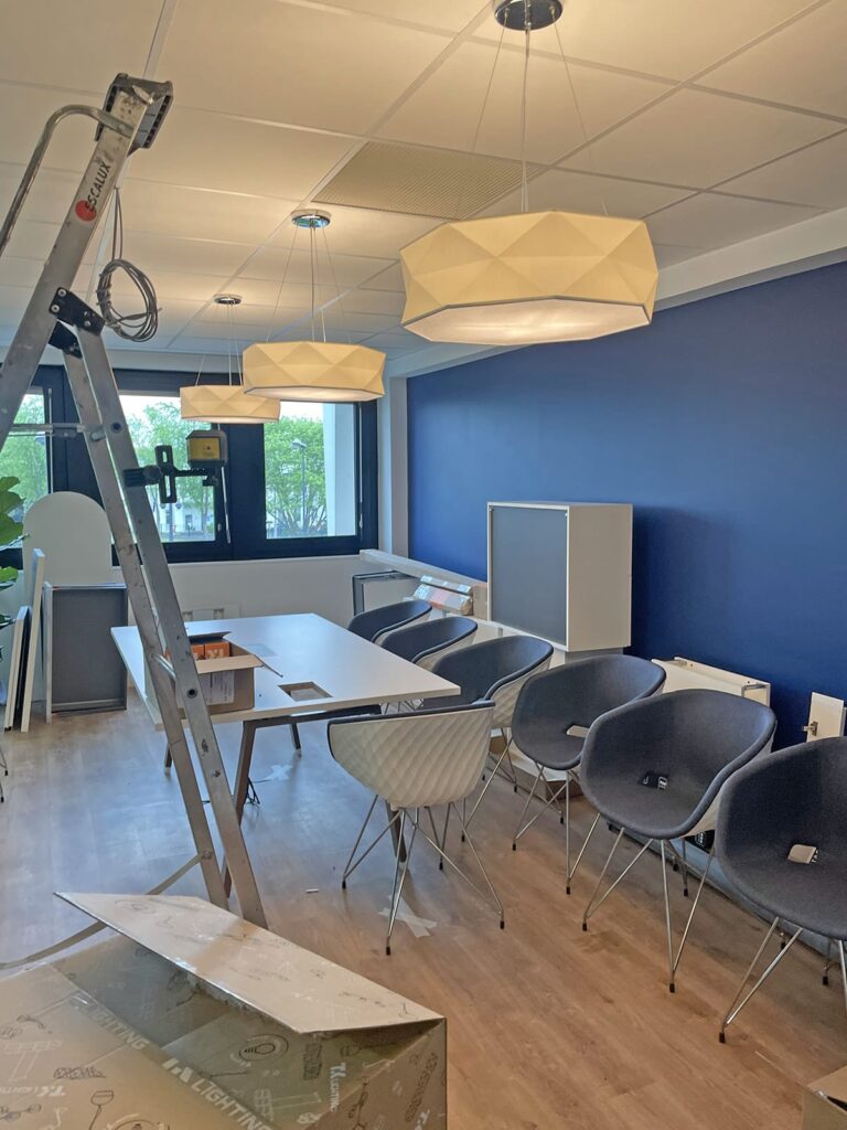 Espace de bureau avec un mur d'accent bleu, des chaises modernes, une table, des lampes suspendues et une échelle, suggérant une installation ou une mise en place en cours.