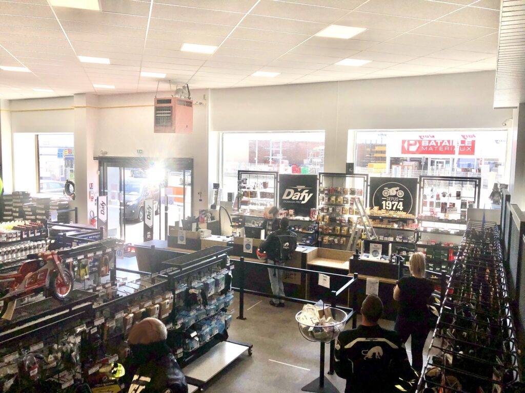 Un magasin d'accessoires pour motos bien éclairé avec des étagères de produits, plusieurs clients qui naviguent, et du personnel derrière la caisse près de grandes fenêtres.
