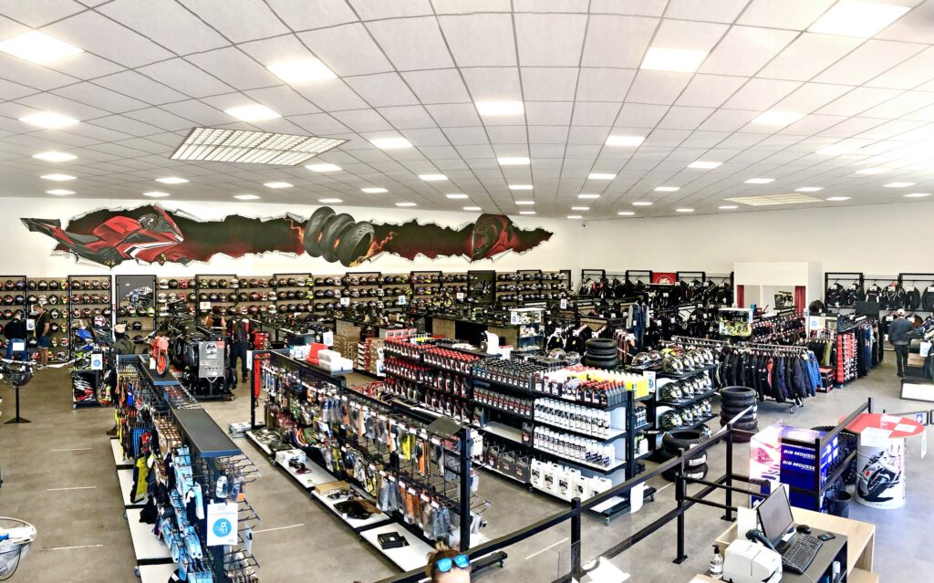 Vue panoramique d'un magasin d'équipement et d'accessoires pour motocycles avec des casques, des vestes, des gants et d'autres équipements soigneusement présentés sur des étagères et des rayonnages.
