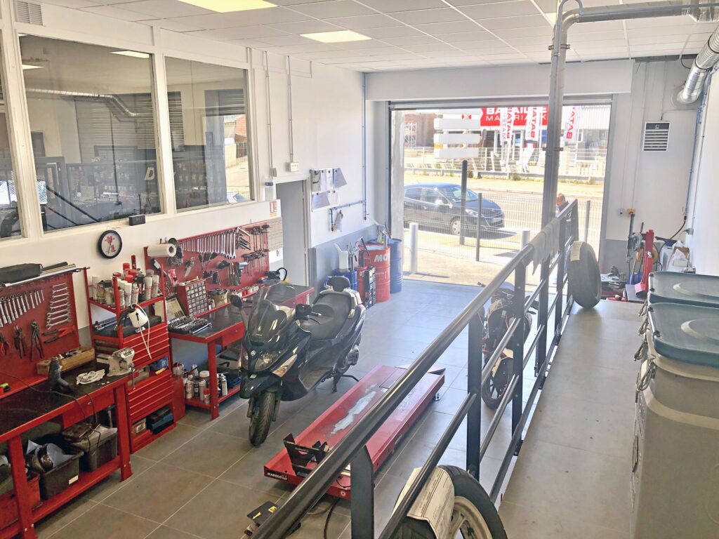 Intérieur d'un atelier de réparation de motos avec deux scooters, des armoires à outils rouges, des établis et une porte de garage ouverte donnant sur l'extérieur.