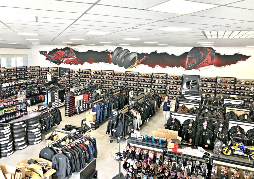Un magasin d'équipement pour motards avec des étagères présentant des casques, des vestes et des accessoires ; des œuvres d'art murales représentant des graphiques sur le thème de la moto décorent le mur supérieur.