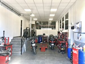 Un atelier de réparation de motos avec des outils, de l'équipement et deux motos placés près d'établis ; des escaliers mènent à une plate-forme surélevée sur la gauche.