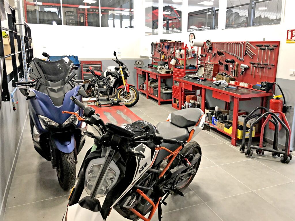 Trois motos sont garées dans un atelier avec des armoires à outils rouges, des établis et divers outils rangés sur les murs.