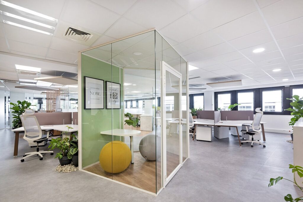Bureau moderne à aire ouverte avec des bureaux blancs, des chaises ergonomiques, des plantes en pot et un module de réunion en verre contenant une petite table ronde et deux sièges sphériques.