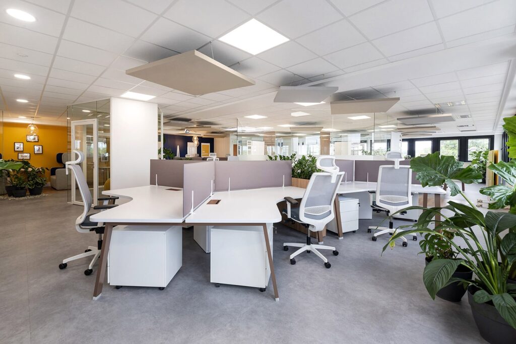 Bureau moderne à aire ouverte avec des bureaux blancs, des chaises ergonomiques, des écrans de séparation, des plantes d'intérieur et de la lumière naturelle provenant de grandes fenêtres. Le bureau est rangé et inoccupé.