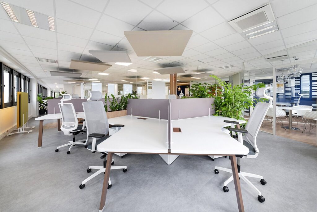 Bureau moderne à aire ouverte avec des bureaux blancs, des chaises ergonomiques, des cloisons de séparation, des plantes en pot et de grandes fenêtres laissant entrer la lumière naturelle.