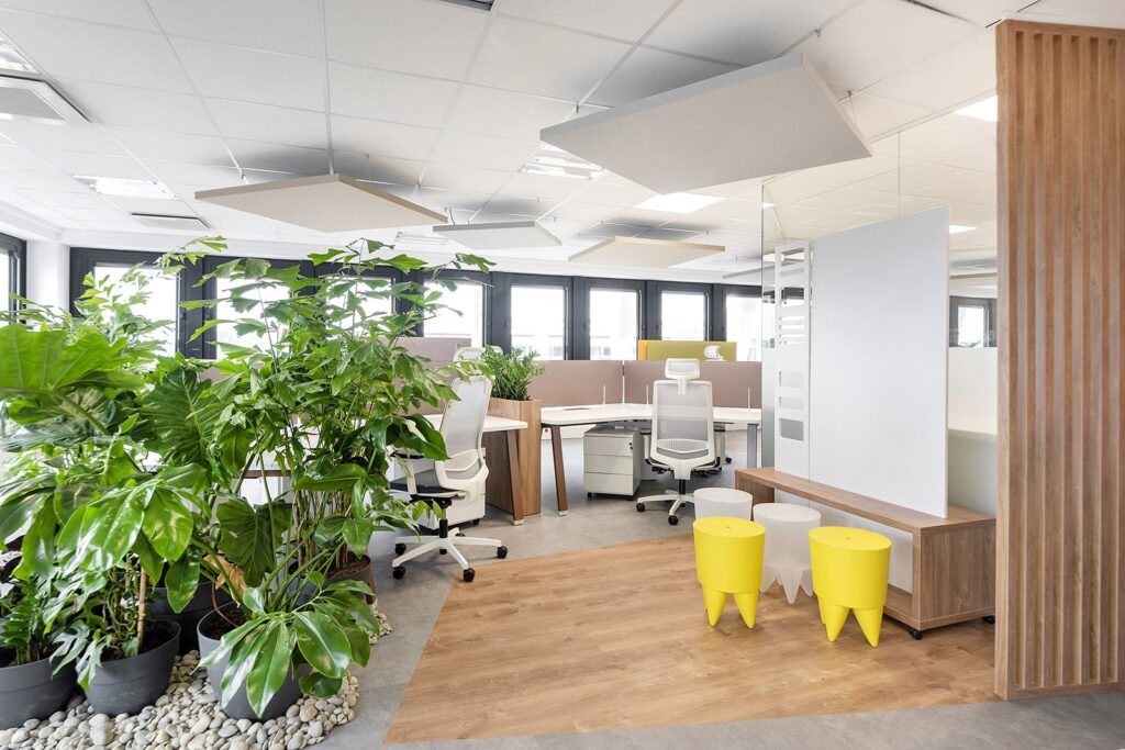 Bureau moderne à aire ouverte avec des bureaux blancs, des chaises ergonomiques, de grandes plantes d'intérieur, trois tabourets jaunes et la lumière naturelle provenant de plusieurs fenêtres.