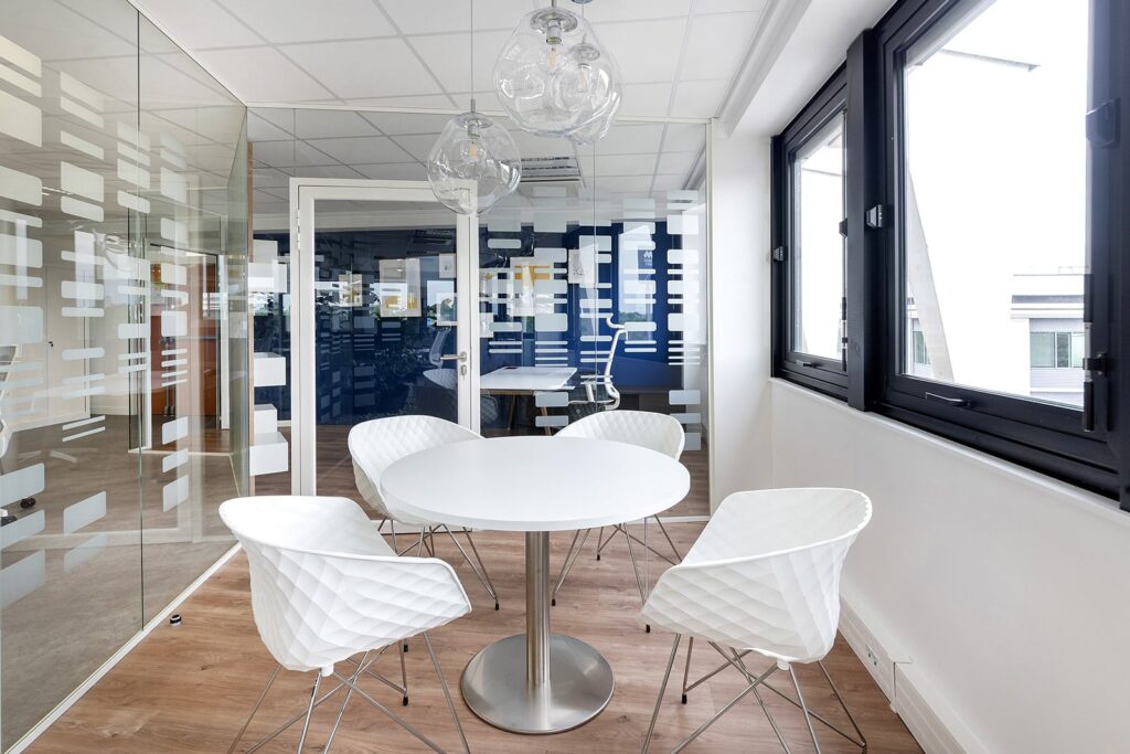 Une salle de réunion moderne avec une table ronde blanche, quatre chaises blanches, des murs en verre avec des motifs géométriques, un sol en bois et de grandes fenêtres.