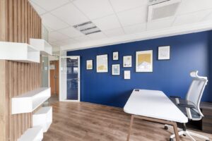 Bureau moderne avec un mur d'accent bleu, des œuvres d'art encadrées, un bureau blanc, une chaise ergonomique, un sol en bois et des étagères. Une porte vitrée mène à une autre pièce.