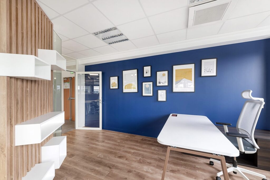 Bureau moderne avec un mur d'accent bleu, une œuvre d'art encadrée, un sol en bois, un bureau blanc et une chaise ergonomique. Des étagères et une porte vitrée sont visibles sur le côté gauche.