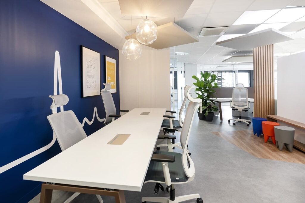Espace de bureau moderne avec des bureaux blancs et des chaises ergonomiques, un mur d'accent bleu, des lampes suspendues, des plantes d'intérieur et la lumière naturelle des grandes fenêtres.