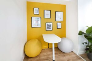 Un petit bureau avec deux chaises à ballon d'exercice est placé contre un mur d'accent jaune décoré d'œuvres d'art encadrées. Une plante est placée à droite du bureau sur un sol en bois.