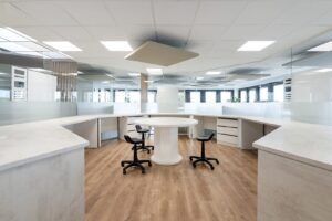 Bureau moderne avec une table ronde blanche, trois chaises pivotantes noires, un sol en bois et des cloisons en verre dépoli sous des plafonniers lumineux.
