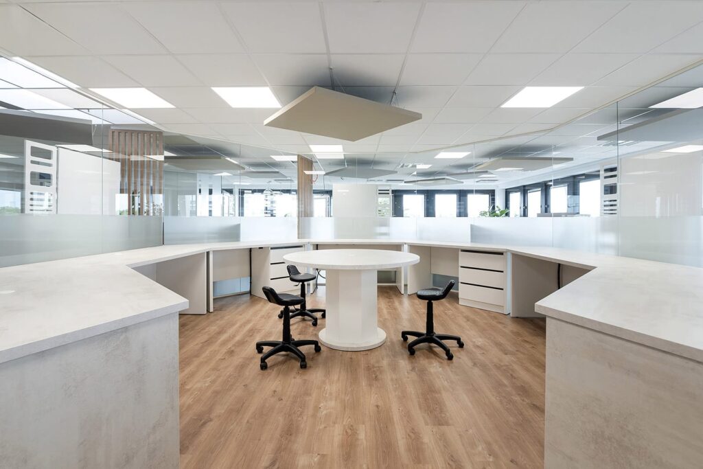 Bureau moderne avec une table ronde blanche, trois chaises de bureau noires, un sol en bois et des cloisons en verre dépoli sous des plafonniers lumineux.