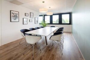 Une salle de conférence moderne avec une table blanche, huit chaises, des œuvres d'art encadrées au mur, un sol en bois, de grandes fenêtres et une plante en pot dans le coin.