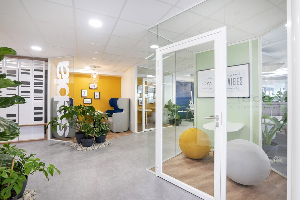Espace de bureau moderne avec une salle de réunion vitrée, des plantes d'intérieur, un coin salon, des œuvres d'art murales motivantes et un éclairage lumineux.