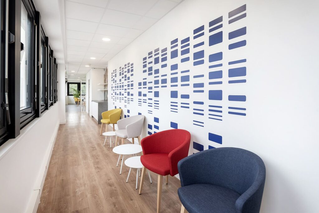 Un couloir au sol en bois est agrémenté de fauteuils colorés et de petites tables rondes le long d'un mur décoré de motifs géométriques bleus.
