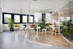 Zone de pause d'un bureau moderne avec des tabourets hauts, de petites tables, des cloisons en verre et plusieurs plantes d'intérieur près de grandes fenêtres.