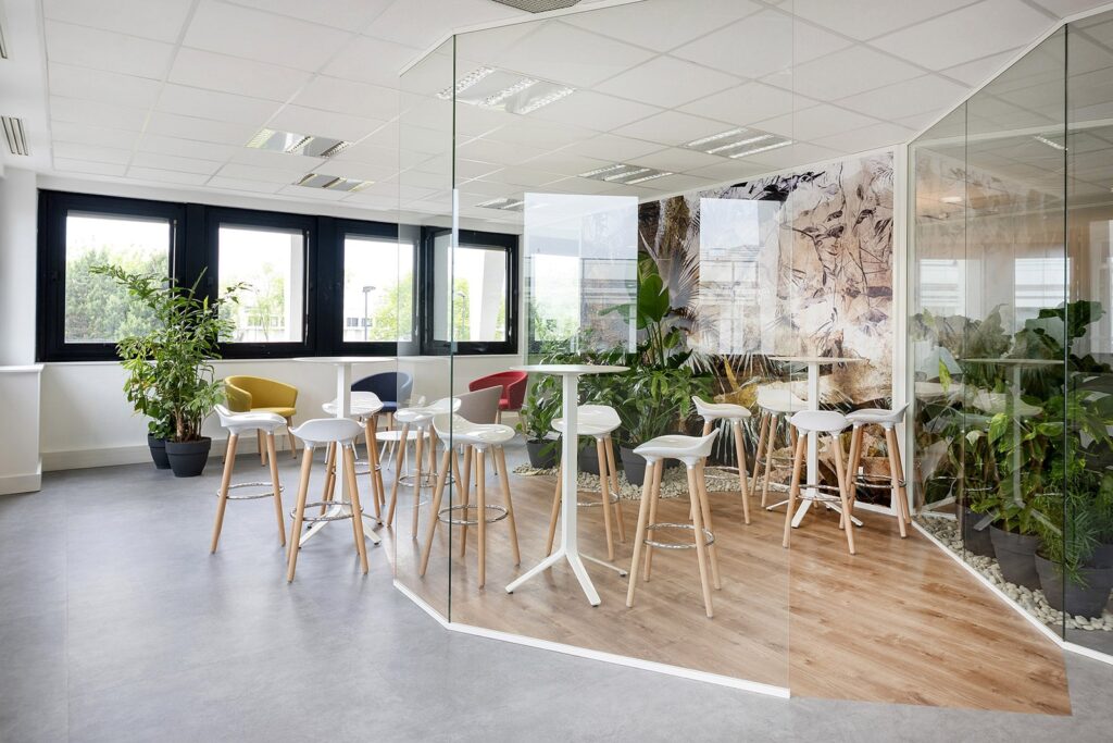 Salon de bureau moderne avec tables hautes, tabourets de bar blancs, cloisons en verre, plantes d'intérieur et grandes fenêtres laissant entrer la lumière naturelle.