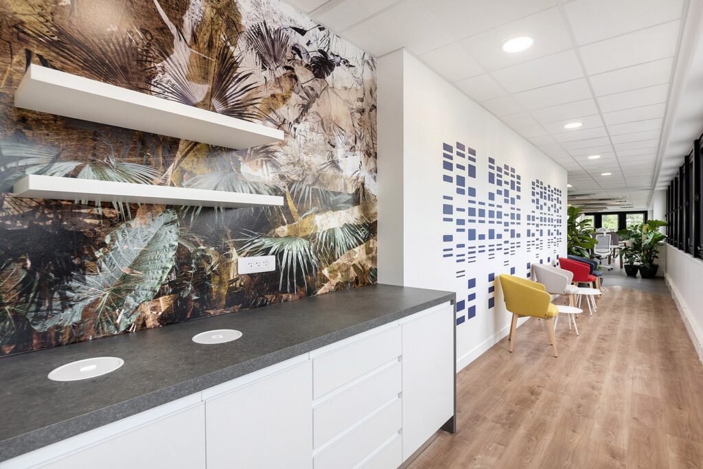 Couloir de bureau moderne avec une kitchenette sur la gauche, une peinture murale abstraite, un sol en bois et des chaises colorées le long du couloir.