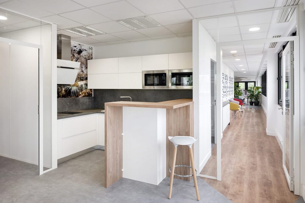 Kitchenette de bureau moderne avec des armoires blanches, des appareils encastrés, un petit bar en bois pour le petit-déjeuner et un tabouret de bar blanc. Un couloir avec des sièges et des plantes est visible à l'arrière-plan.
