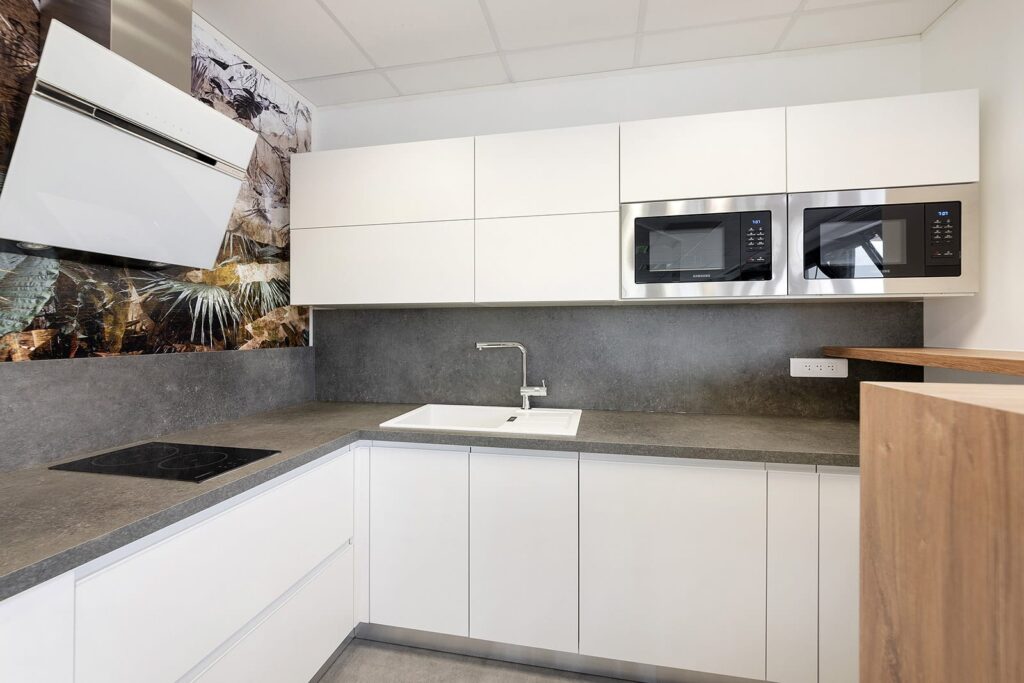 Cuisine moderne avec des armoires blanches, des comptoirs gris, un évier encastré, deux micro-ondes muraux et une table de cuisson, avec un dosseret décoratif sur un mur.