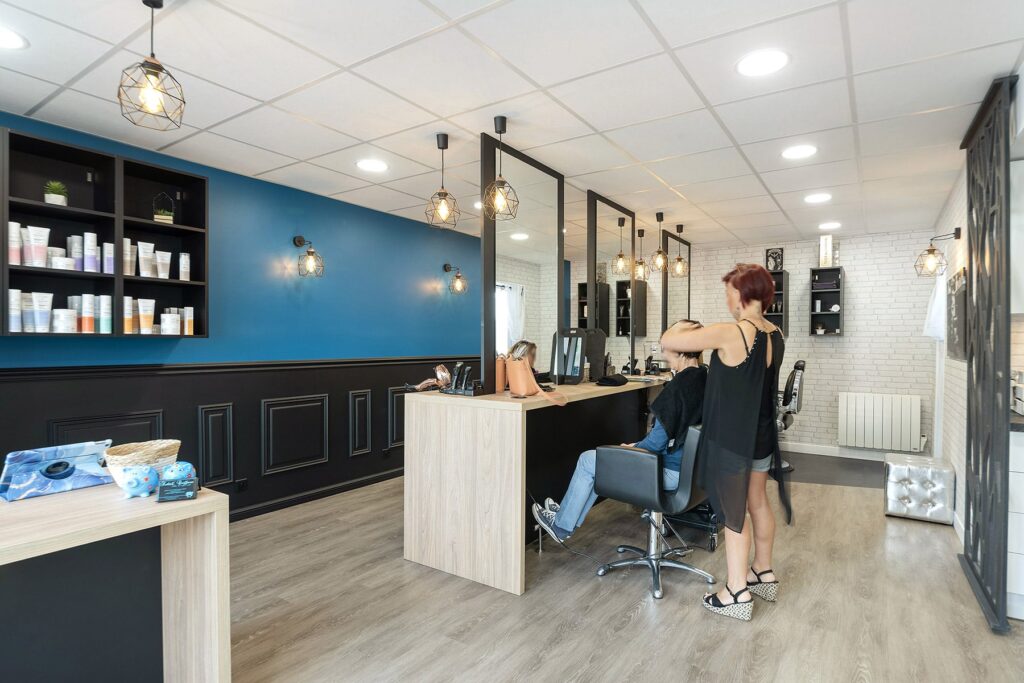 Un salon de coiffure moderne avec un mur d'accent bleu, des étagères de produits capillaires et un styliste qui coupe les cheveux d'une cliente à un poste de coiffure.